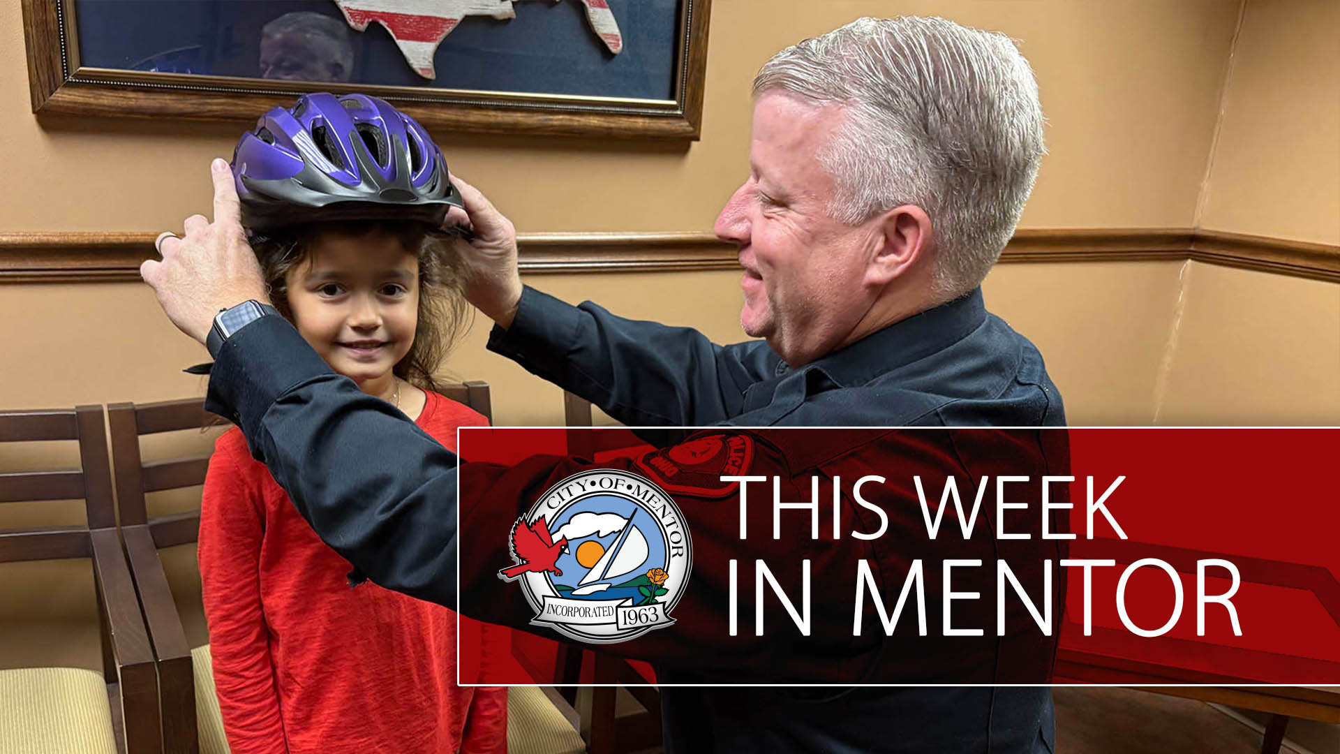 An officer affixing a bicycle helmet on a child with the words "This Week in Mentor" overlayed in a semitransparent red field.